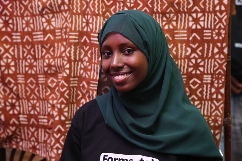 A smiling young woman in a green headscarf and a Kabakoo shirt, in front of a bogolan cloth.