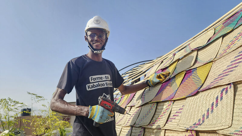 A builder in a hard hat fixes handwoven fan panels onto the roof structure of Le Nid de Bamako.